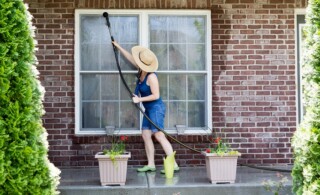 Housewife standing on a patio washing the windows of her house with a hose attachment as she spring-cleans the exterior at the start of the new summer season