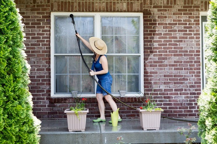 Housewife standing on a patio washing the windows of her house with a hose attachment as she spring-cleans the exterior at the start of the new summer season