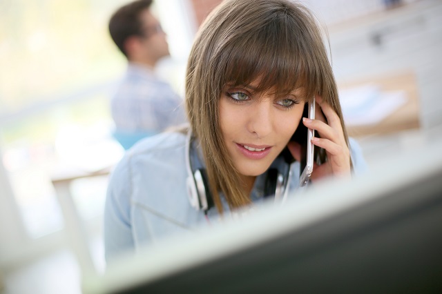 Young woman talking on smartphone in front of desktop.