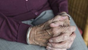 An older gentleman sits in his home with his hands clasped.