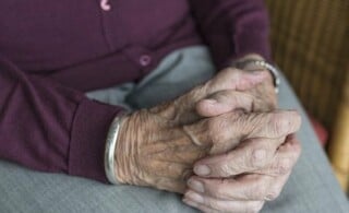 An older gentleman sits in his home with his hands clasped.