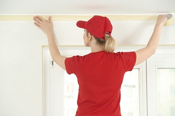 Woman painter worker protecting ceiling moulding with masking tape before painting at home improvement work