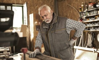 Mature carpenter experiencing back pain while checking wood plank in his workshop