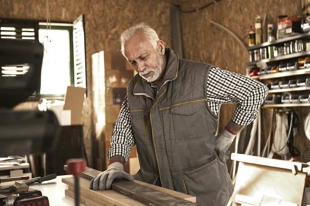 Mature carpenter experiencing back pain while checking wood plank in his workshop