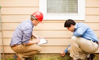 Repairmen, building inspectors, exterminators, engineers, insurance adjusters, or other blue collar workers examine a building/home's exterior wall and foundation. One wears a red hard hat and clear safety glasses and holds a clipboard. The other checks the foundation with tool.