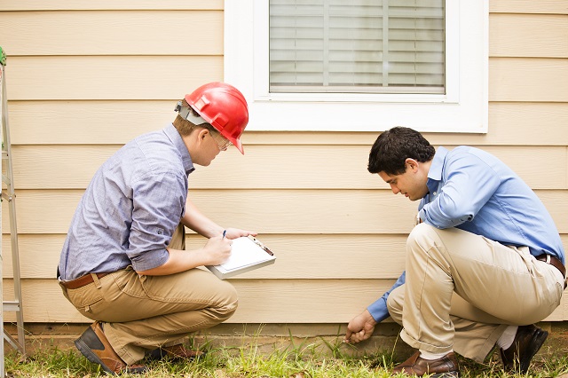 Repairmen, building inspectors, exterminators, engineers, insurance adjusters, or other blue collar workers examine a building/home's exterior wall and foundation. One wears a red hard hat and clear safety glasses and holds a clipboard. The other checks the foundation with tool.