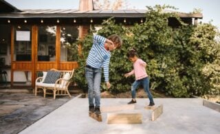 Two children balance on wooden blanks on concrete driveway