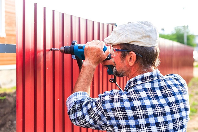 Man in plaid shirt and hat screws a vinyl fence board into a post