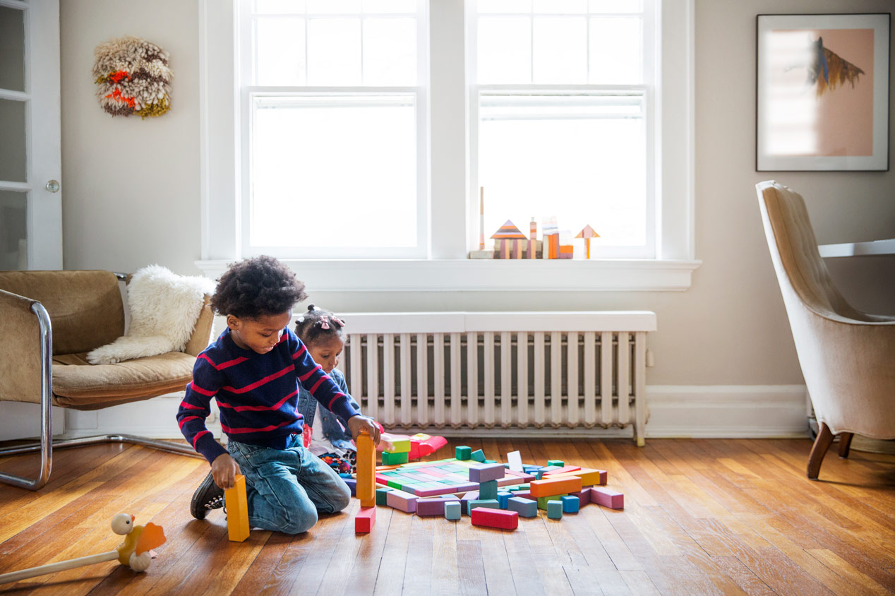 Two children play on wooden floor with colorful toys in front of white radiator