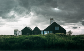 A large house across a grassy field with dark grey clouds casting shadows overhead